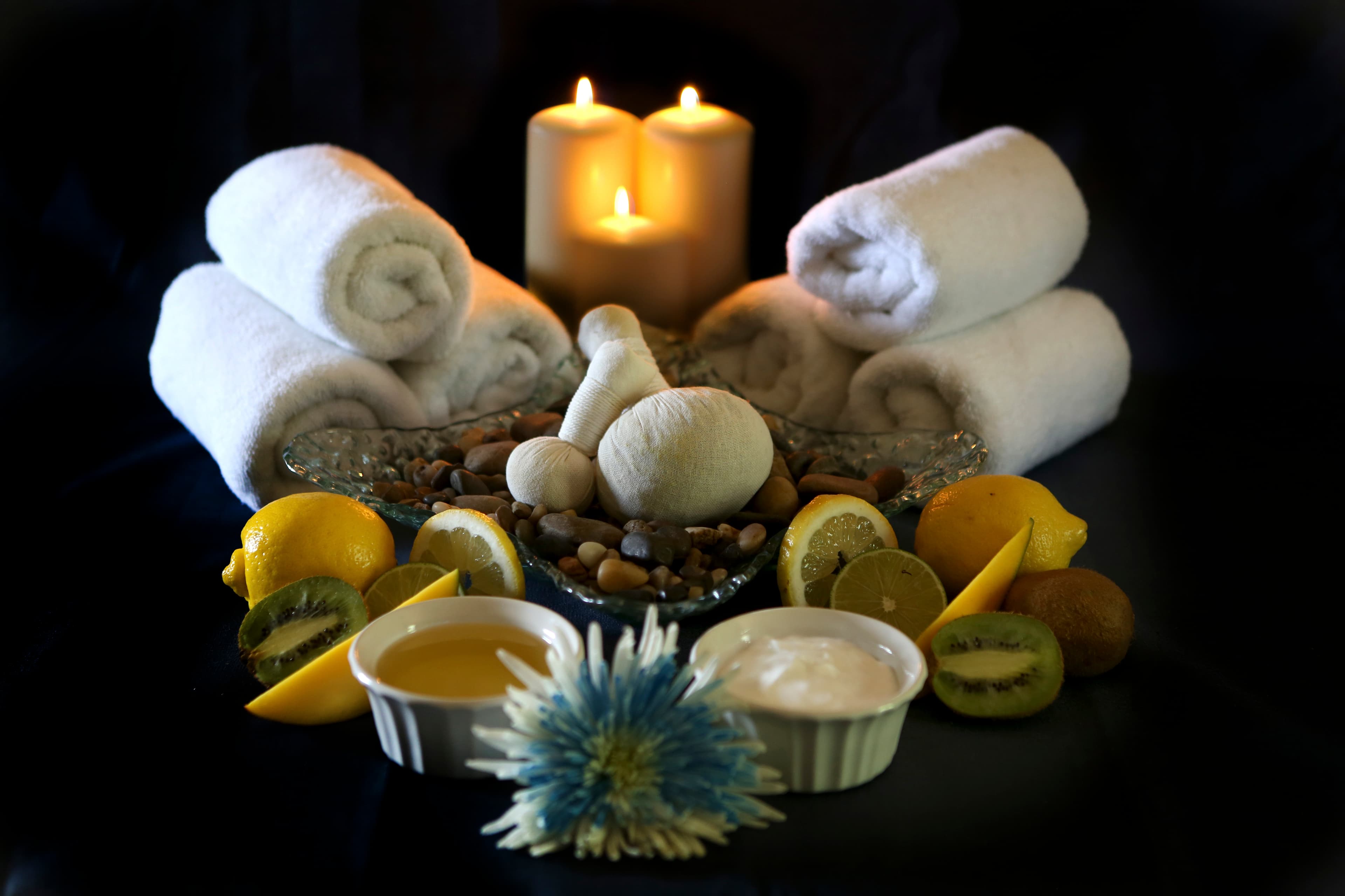 A spa still life with rolled towels, candles, an herbal compress ball, and citrus fruit